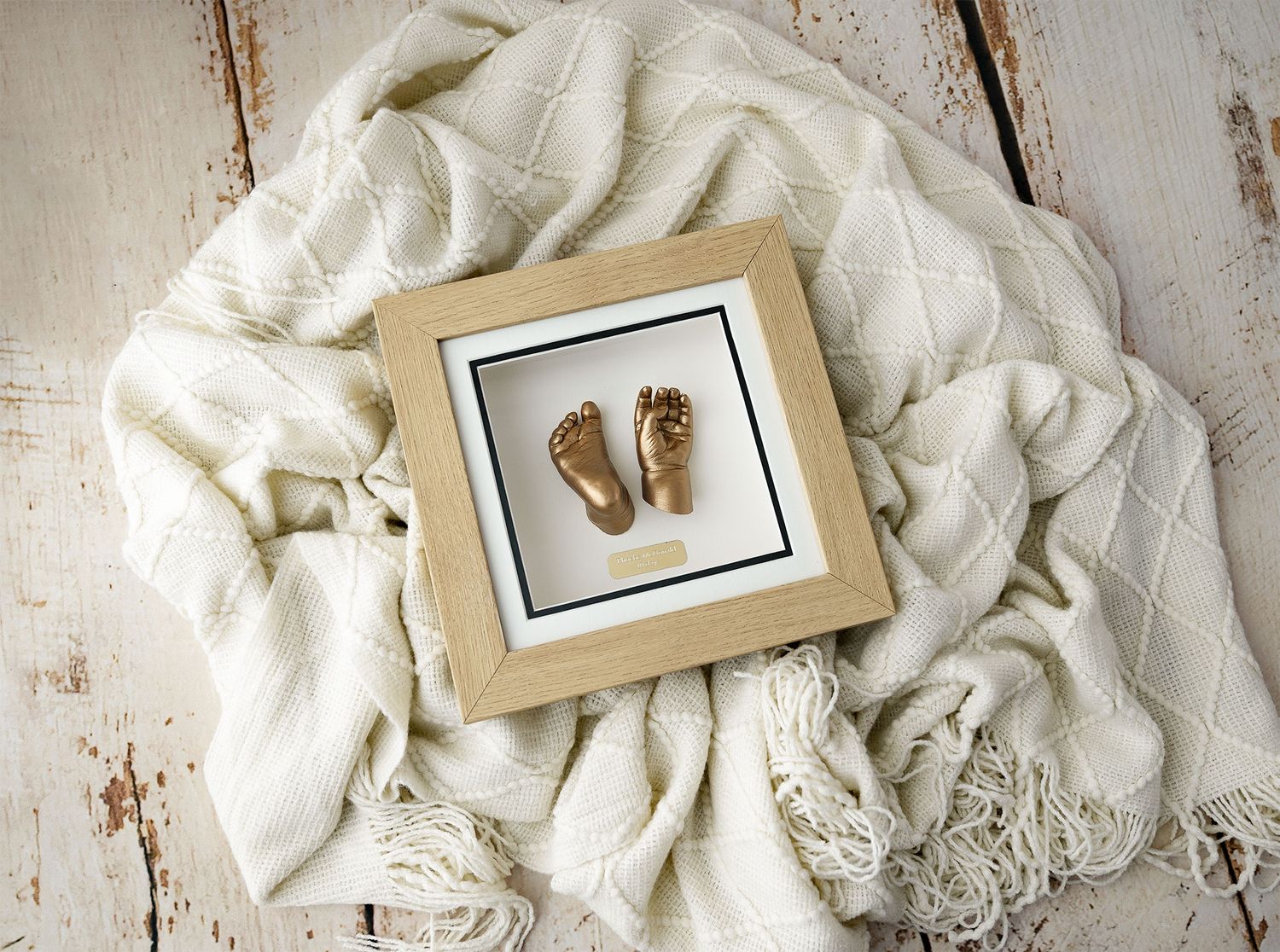 Gold baby hand and foot cast display in black shadow box frame, with white floral arrangement on woven mat. Newborn photographer Argyll