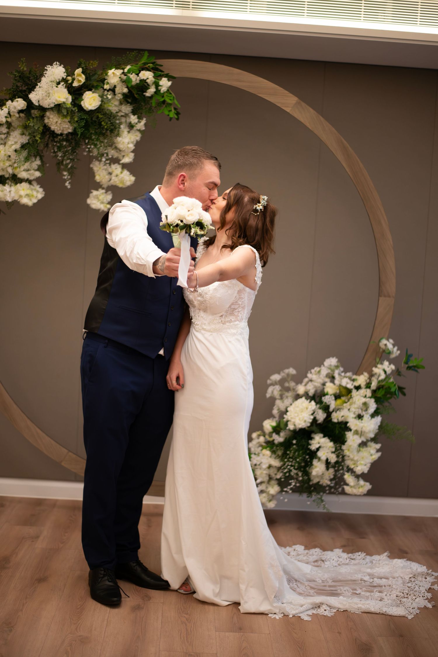 Bride and groom share a kiss at indoor wedding ceremony in front of circular floral arch with white flowers.