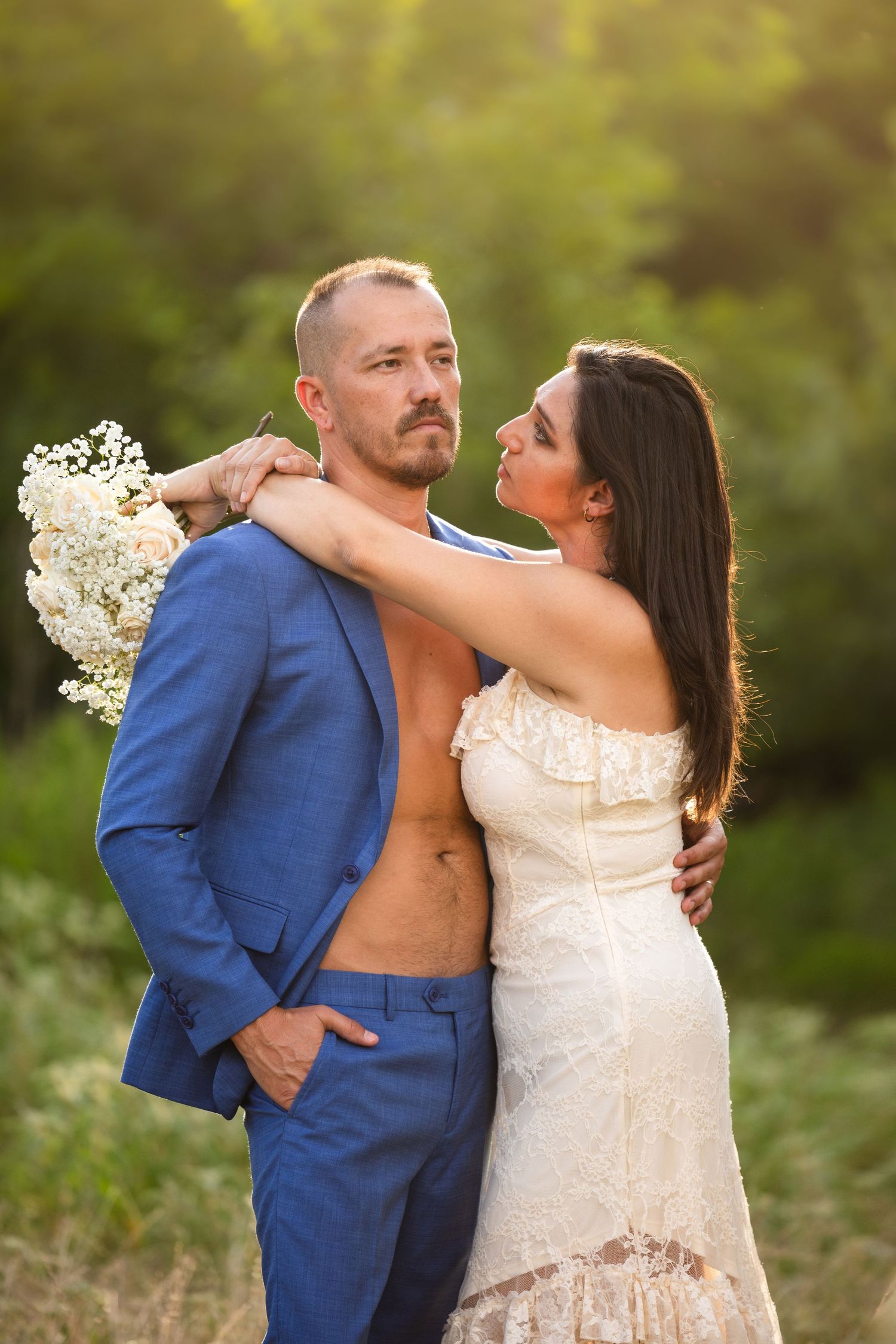 A couple poses in a field, man in open blue suit and woman in white lace dress holding a bouquet, with green trees behind.