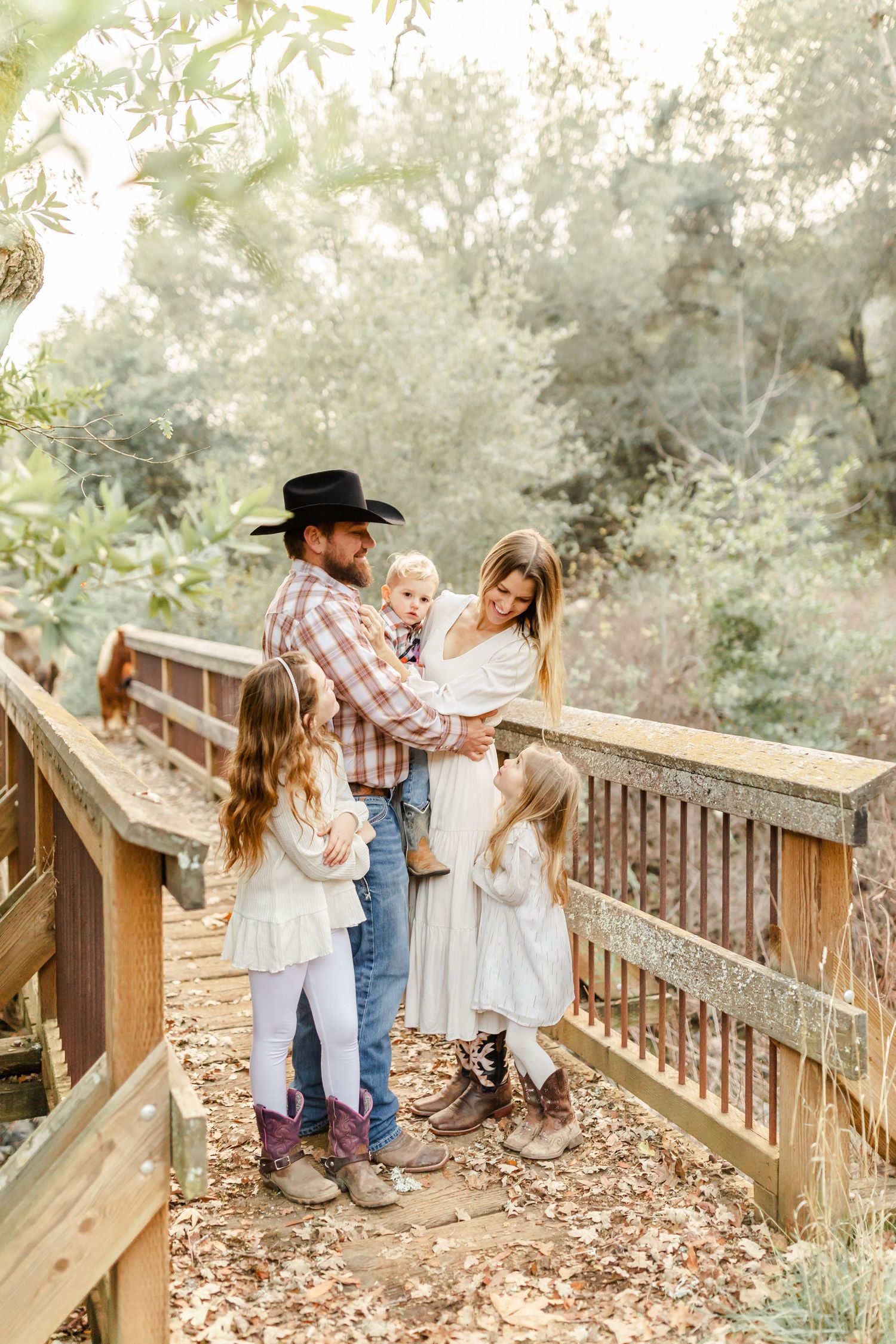 Bearded cowboy in black hat holds toddler in matching plaid shirts and jeans outdoors near cattle.