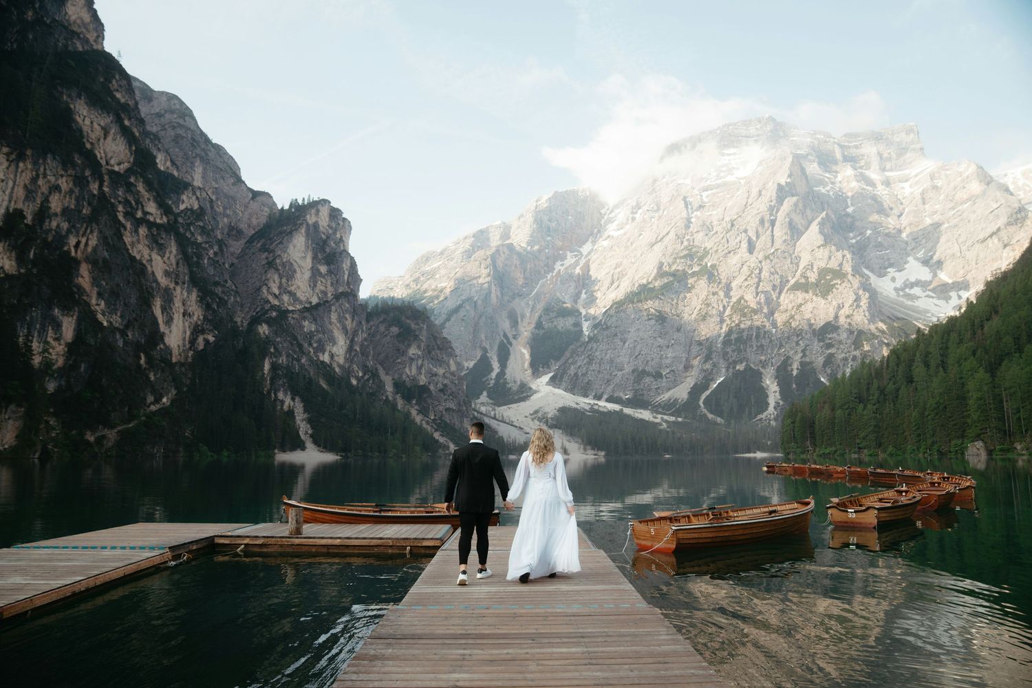 Bride and groom walk hand-in-hand on a wooden dock at Lago di Braies, surrounded by mountains and boats.