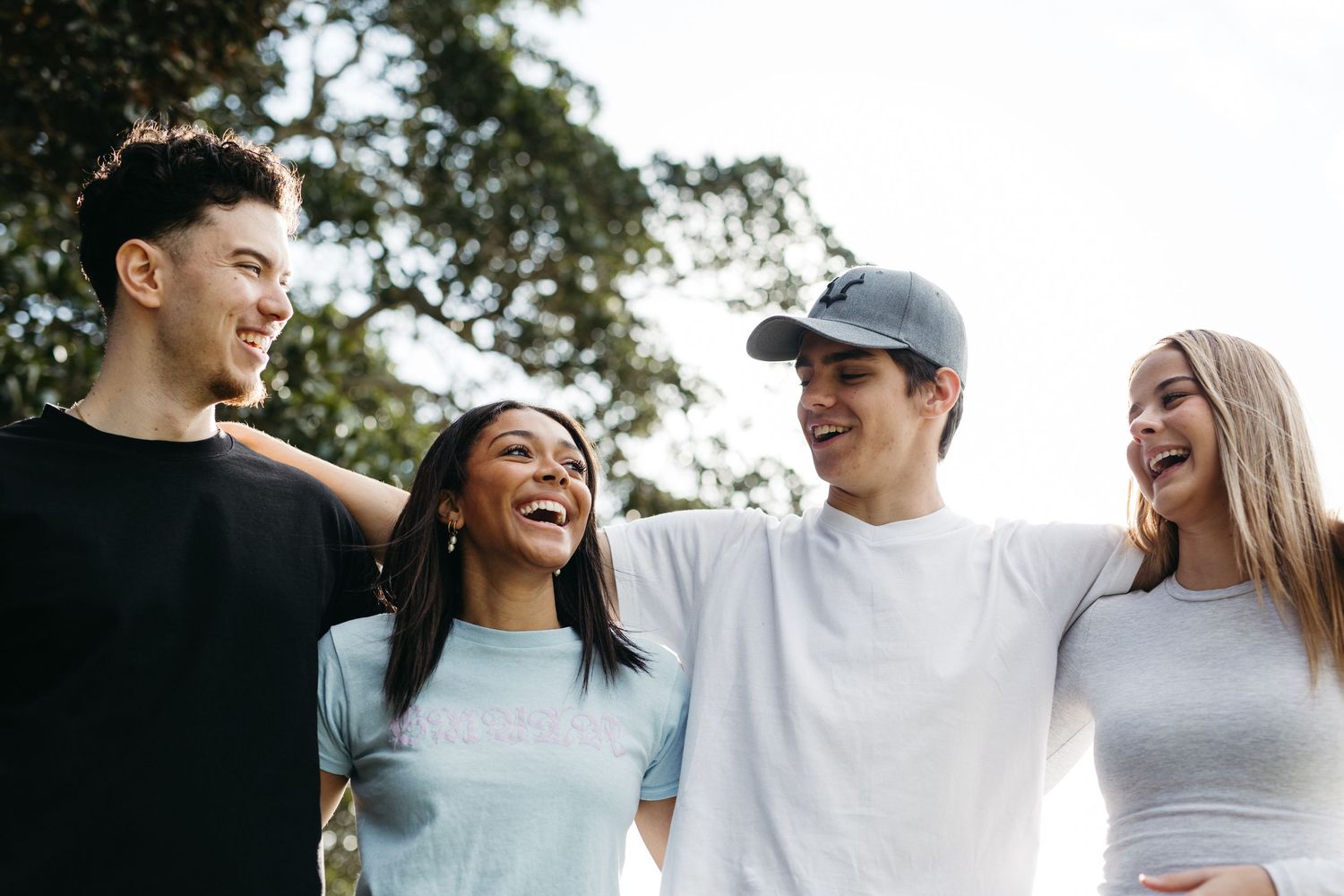 Four happy teenagers laughing and smiling together outdoors, standing close with arms around each other.