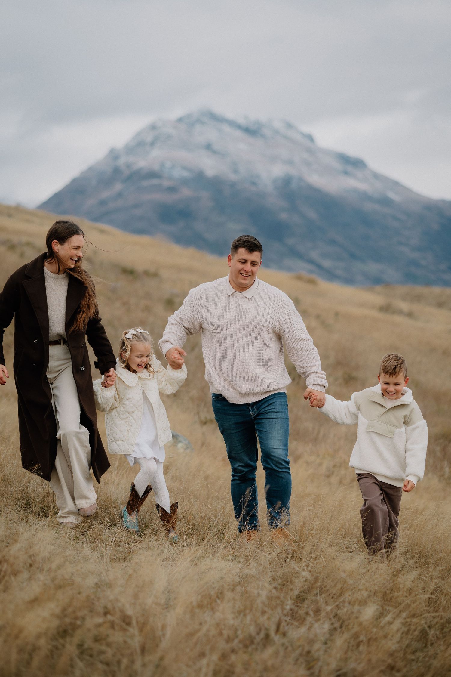 Family of four walking through golden grass fields with snow-capped mountain backdrop on cloudy day.
