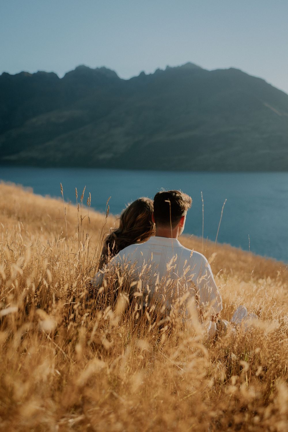 A couple sits in tall golden grass overlooking a mountain lake on a sunny afternoon.