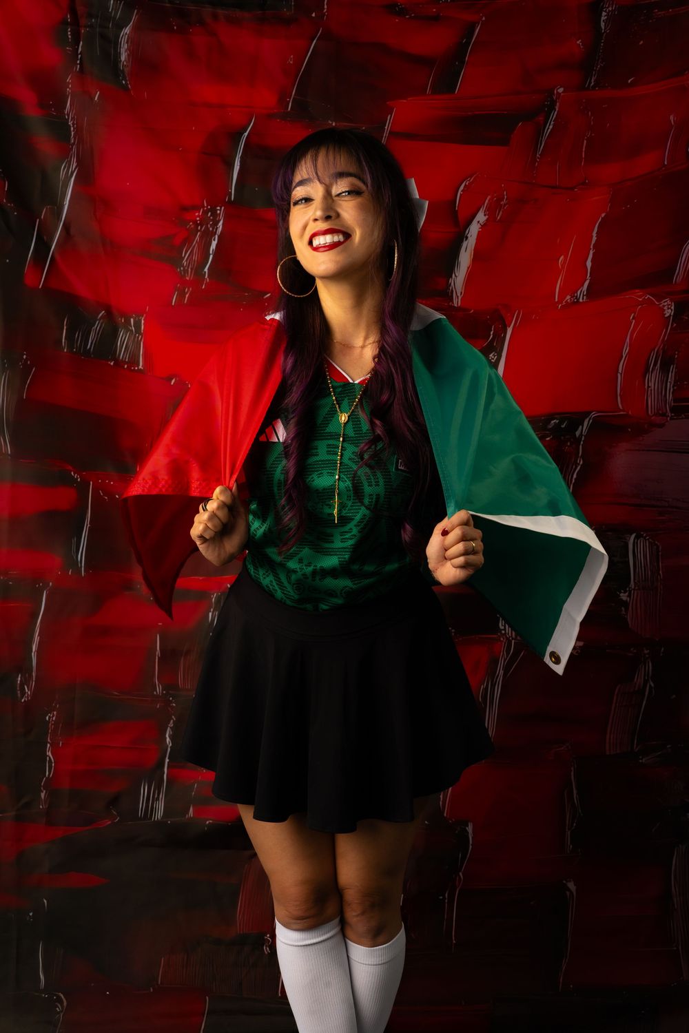 A young woman smiles holding a Mexican flag, wearing a green jersey and black skirt against a red textured background.