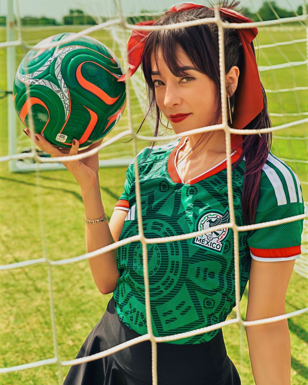 Woman in Mexico soccer jersey holds a football against a goal net on a green field.