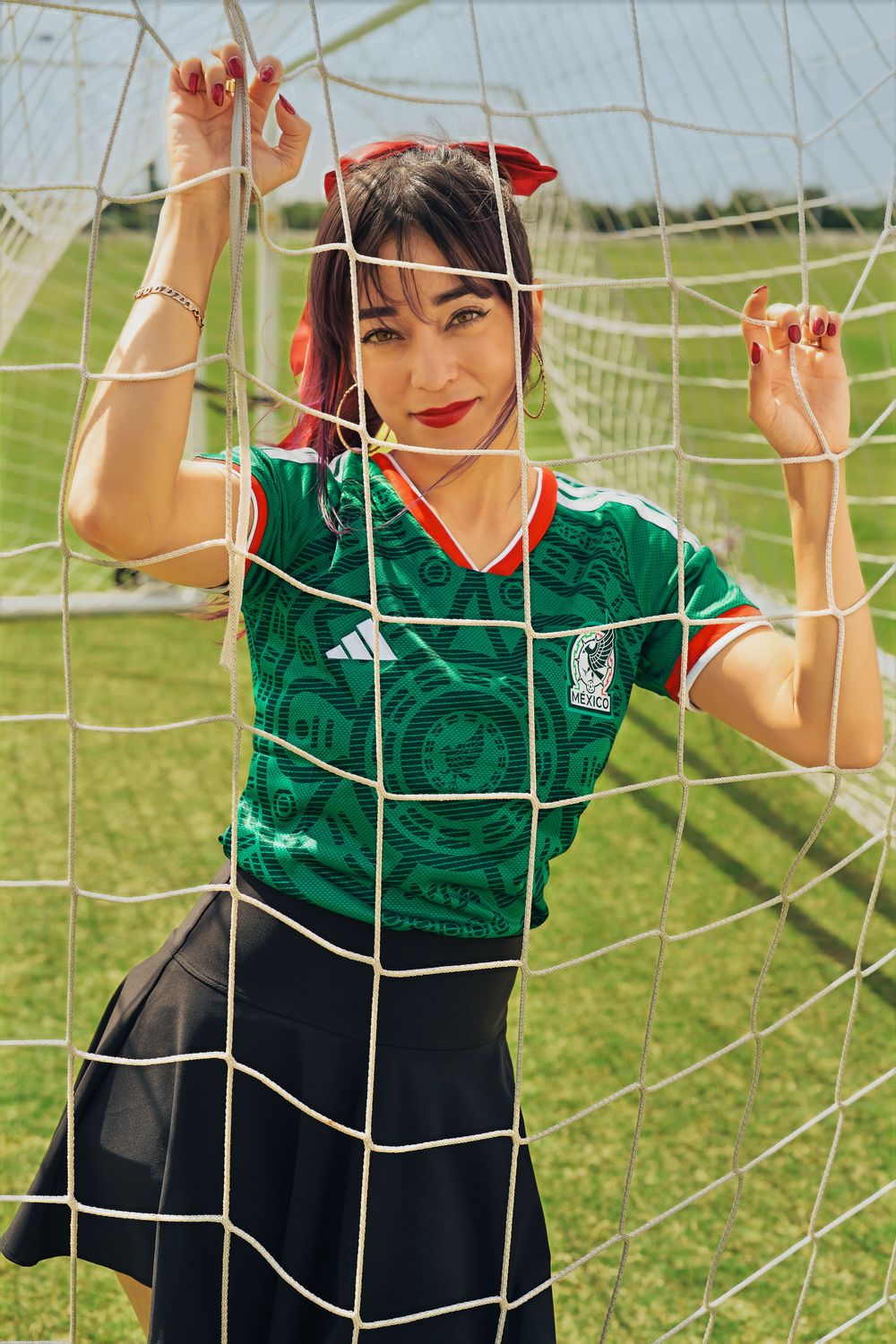 Woman in Mexico soccer jersey poses at goal net on green field, wearing black skirt and red hair bow.