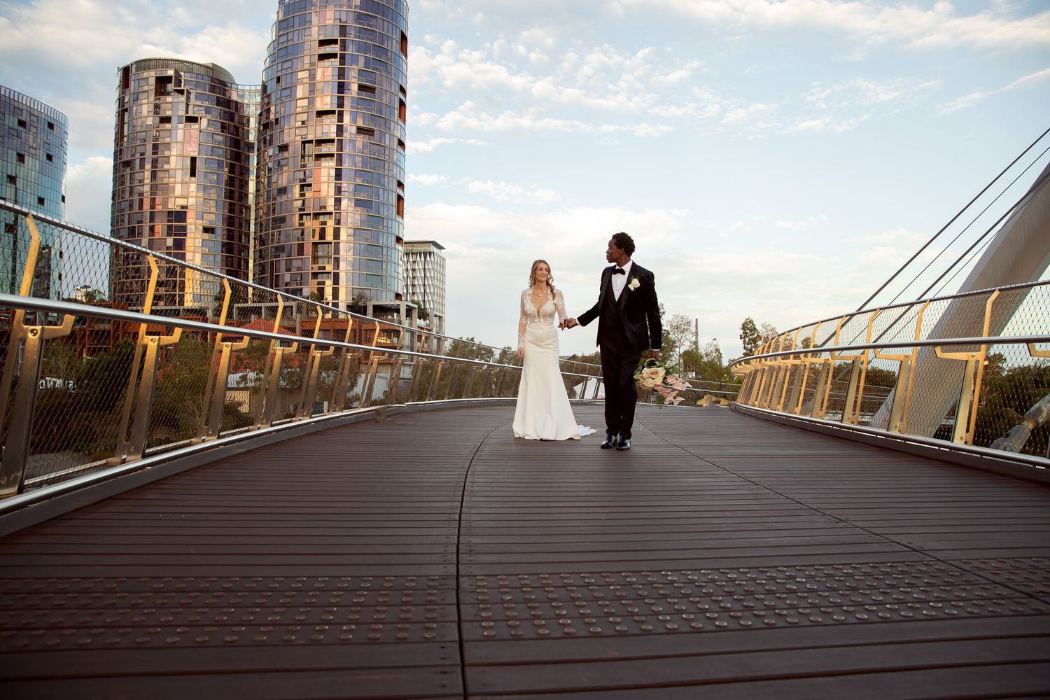 Bride and groom holding hands on a modern bridge with city skyscrapers in the background on their wedding day.