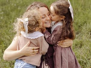 A smiling woman receives kisses on both cheeks from two young girls in a sunny green field.