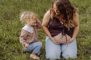 Pregnant woman sitting on grass smiling at toddler girl during outdoor maternity photo session.