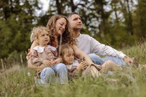 Happy family with two young children sitting together in a lush green meadow surrounded by tall trees.