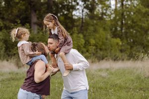Family of four enjoying outdoor fun, parents carrying young daughters on shoulders in a lush green park setting.
