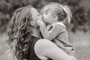 Black and white photo of a mother holding her toddler daughter, sharing a tender kiss outdoors.