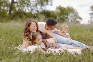 Happy family of four sitting on a blanket in a green meadow, parents cuddling two young children outdoors.
