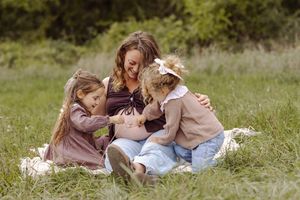 Pregnant woman sitting in grass with two young daughters touching her baby bump during outdoor maternity photo session.