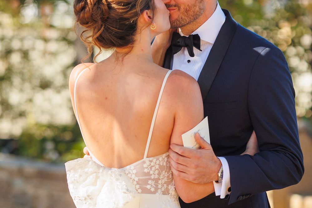Bride and groom sharing a romantic kiss outdoors, wearing elegant wedding attire with lace gown and navy tuxedo.