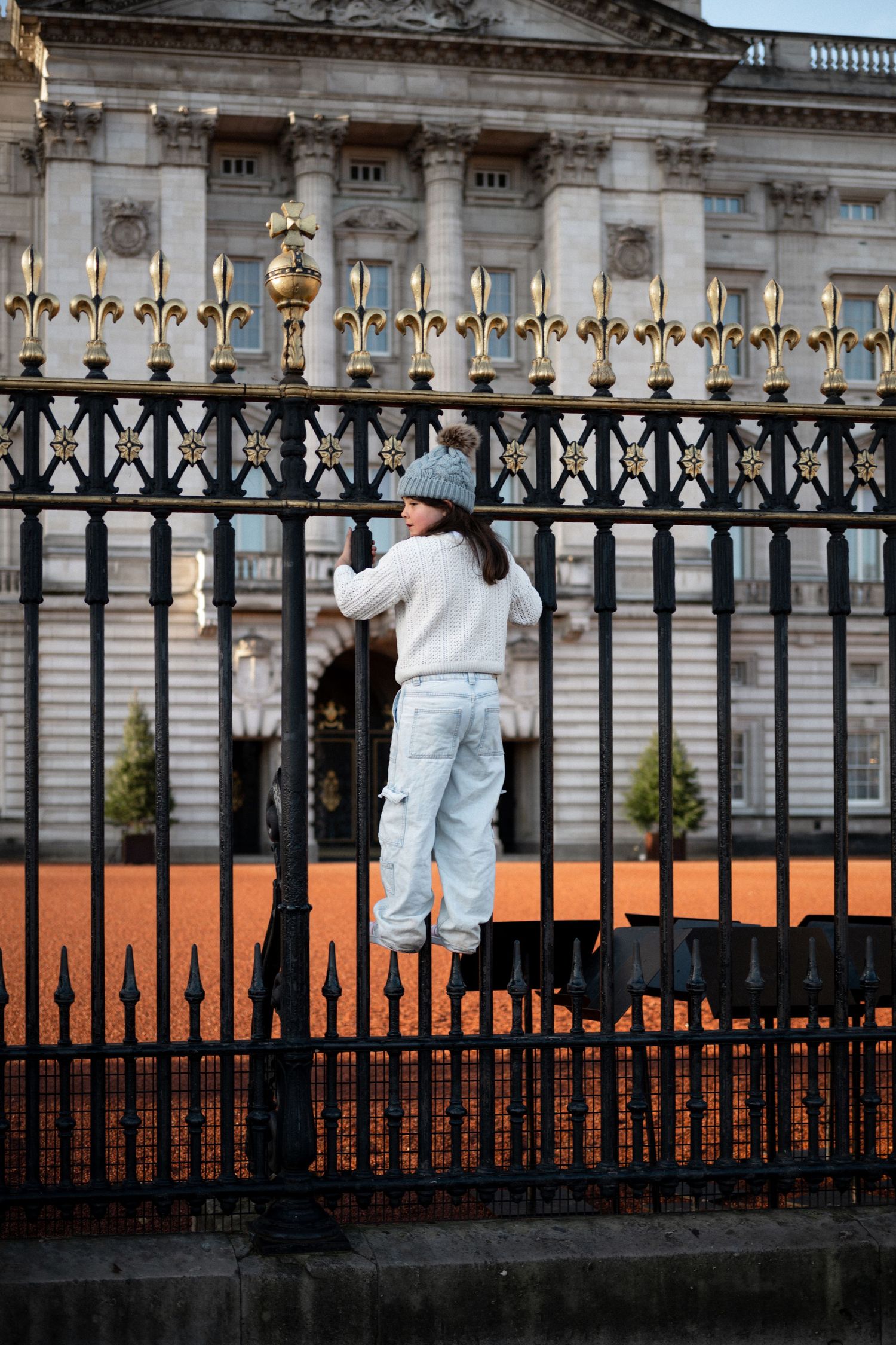 Young woman in white sweater and gray beanie climbs ornate black iron gates at Buckingham Palace, London.