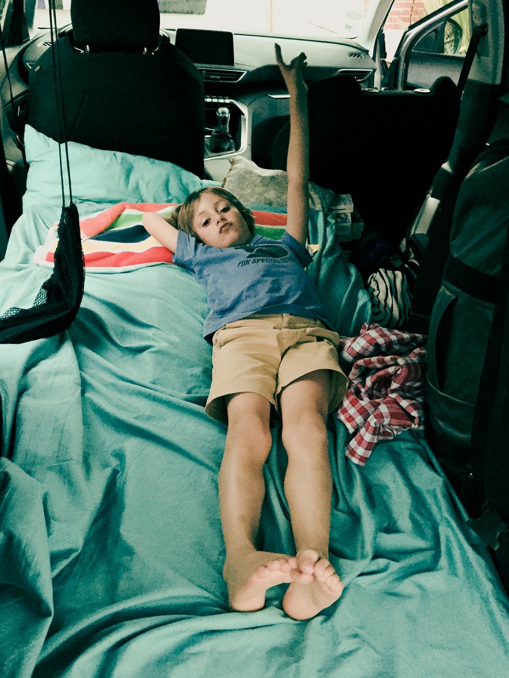 A child lying stretched out on a teal bedding setup inside the back of an SUV, arms raised overhead.