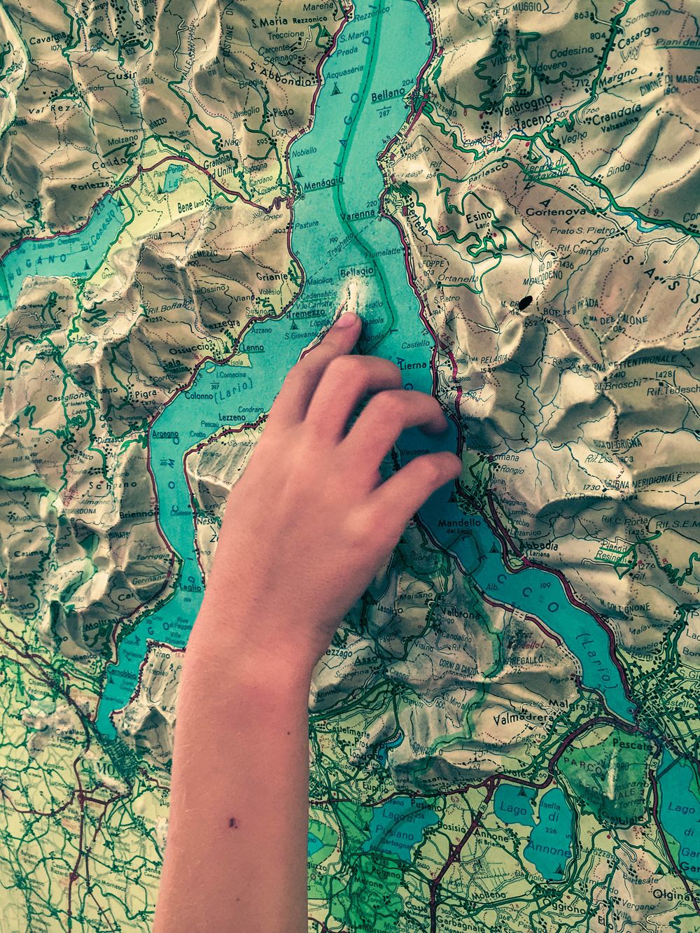 A hand pointing to a location on a detailed topographic map showing a turquoise lake and surrounding terrain.