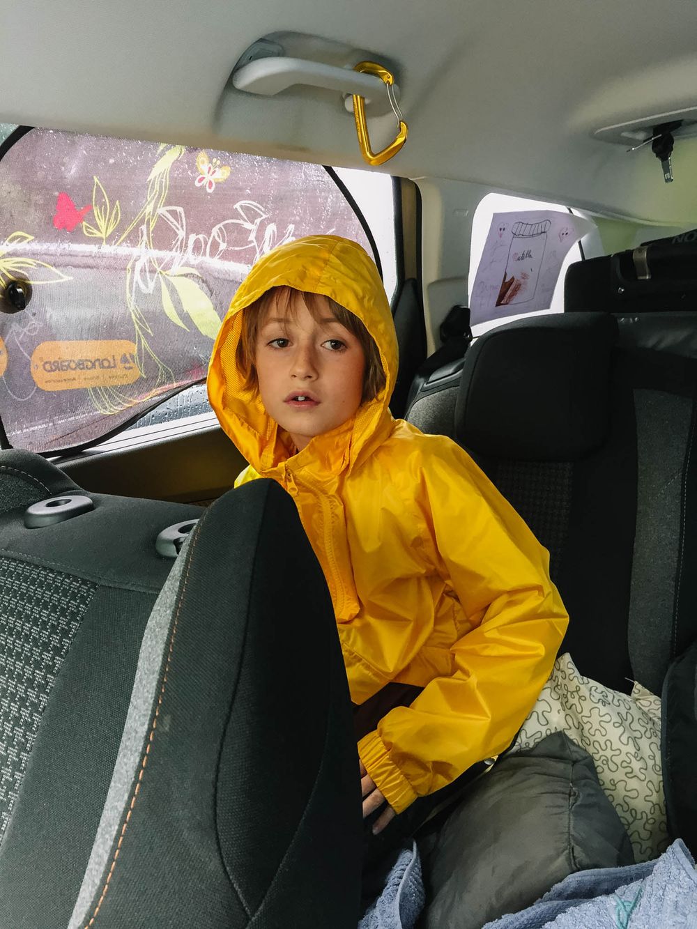 A young child wearing a bright yellow hooded raincoat sits in the back seat of a car, looking over their shoulder.