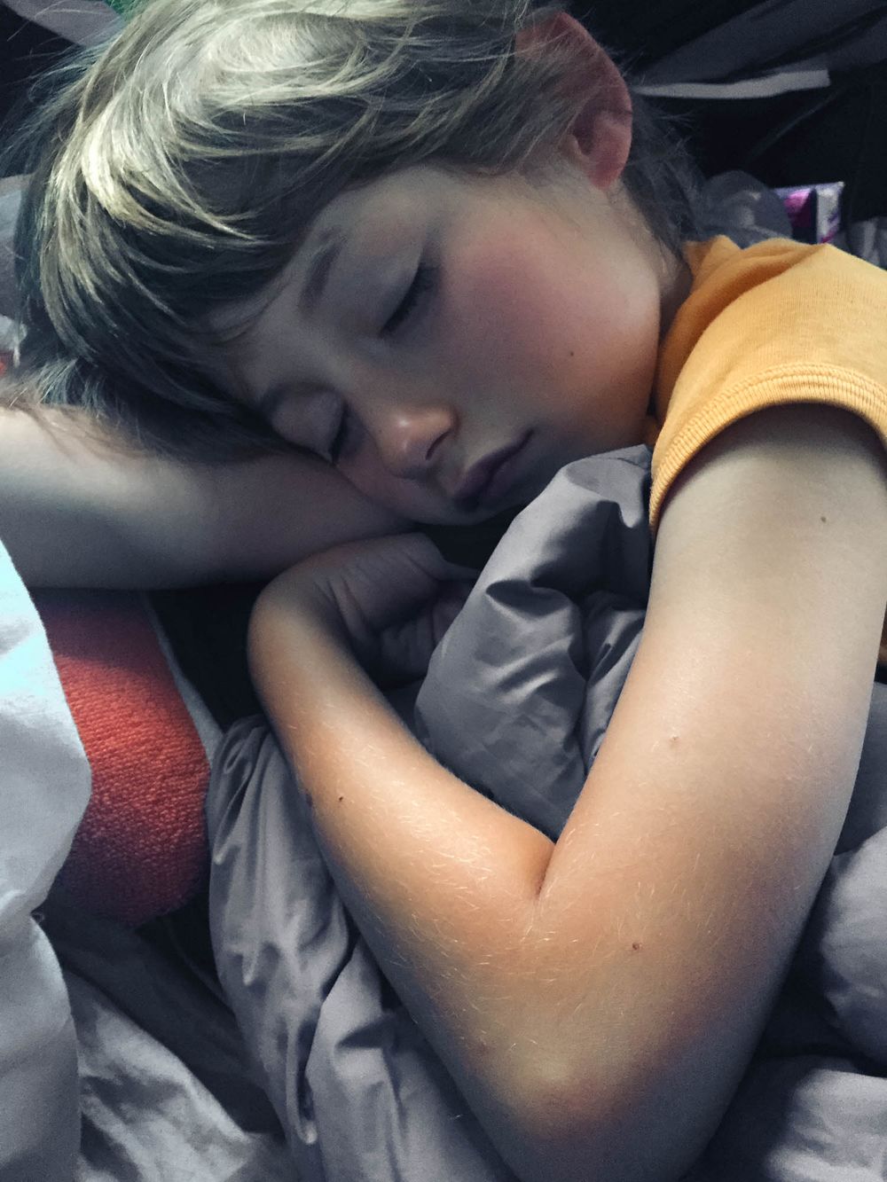 Young child sleeping peacefully on gray bedding, wearing an orange shirt, with rosy cheeks and blonde hair.