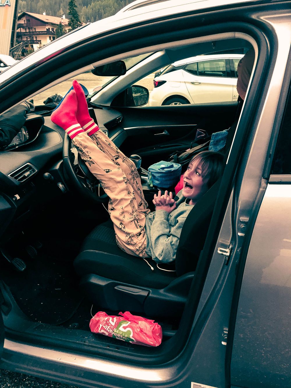 Child lying sideways in car seat with red socks up, laughing with door open in snowy mountain parking lot.