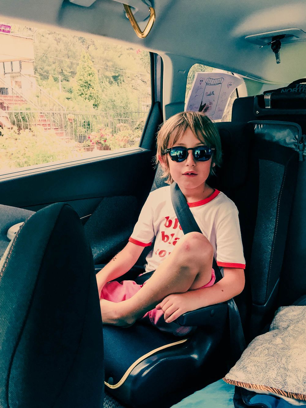 A young child wearing sunglasses and a white t-shirt sits buckled in a car seat inside a vehicle.