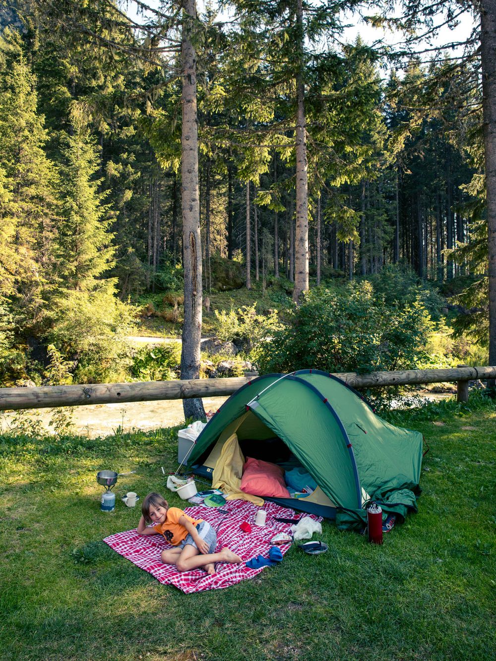 Person relaxing on a blanket beside a green camping tent in a sunny forest clearing near a stream.