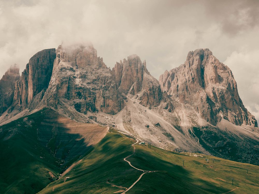 Dramatic Dolomite mountain peaks rise above lush green valleys under moody overcast skies in the Italian Alps.