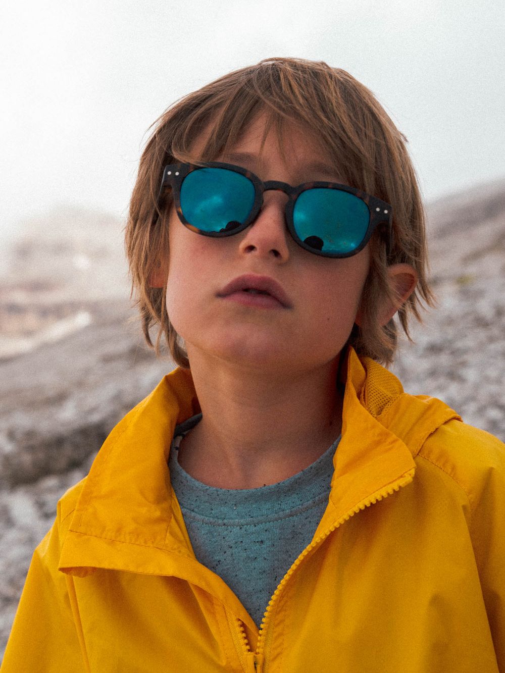 Young boy wearing blue mirrored round sunglasses and yellow jacket outdoors.