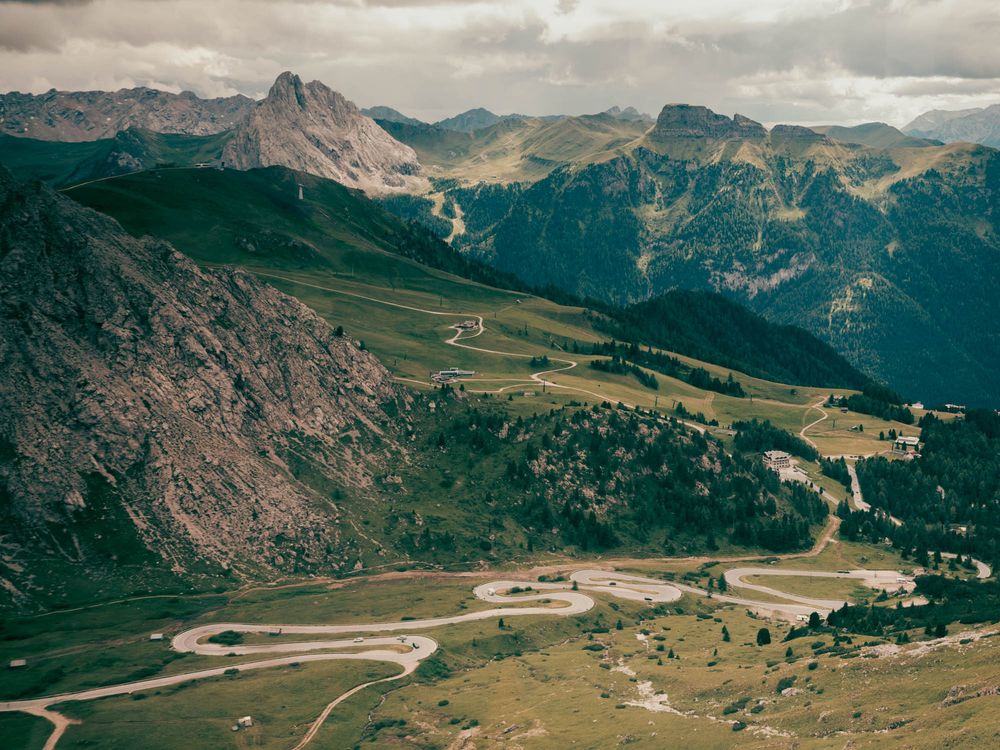 Aerial view of winding mountain road switchbacks through lush green valleys in the Dolomites, Italy.