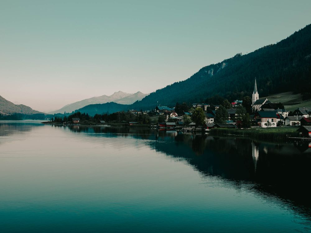 A serene alpine lake reflects a charming village with a church steeple, surrounded by lush mountains under a clear sky.