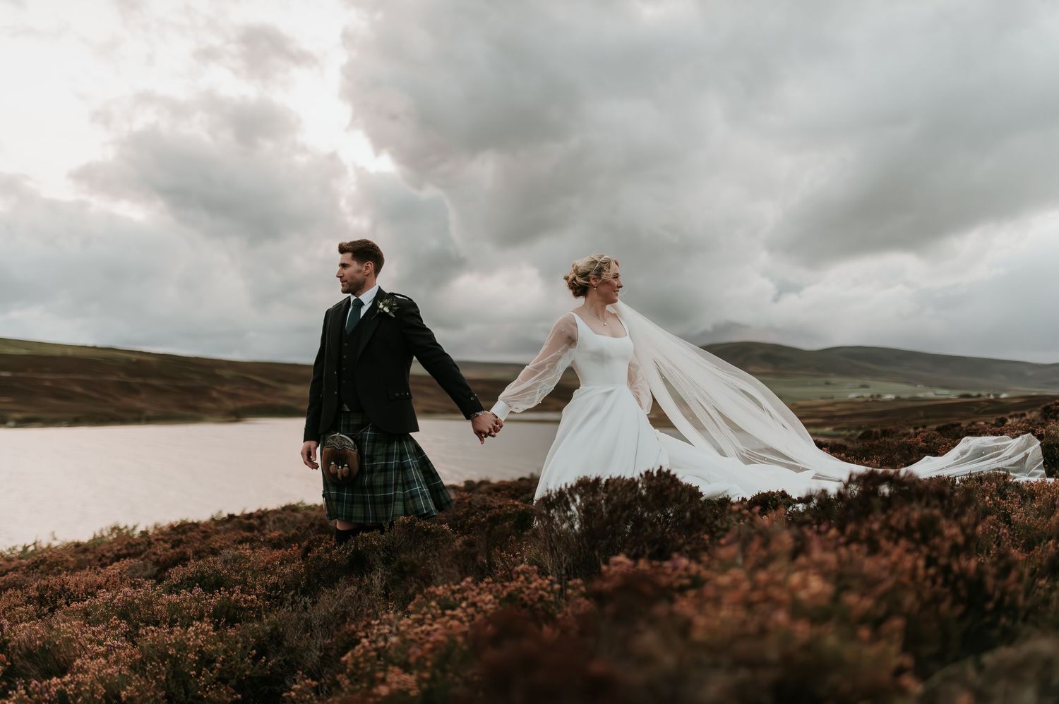 A couple in wedding attire holding hands against dramatic cloudy skies in the Scottish Highlands.