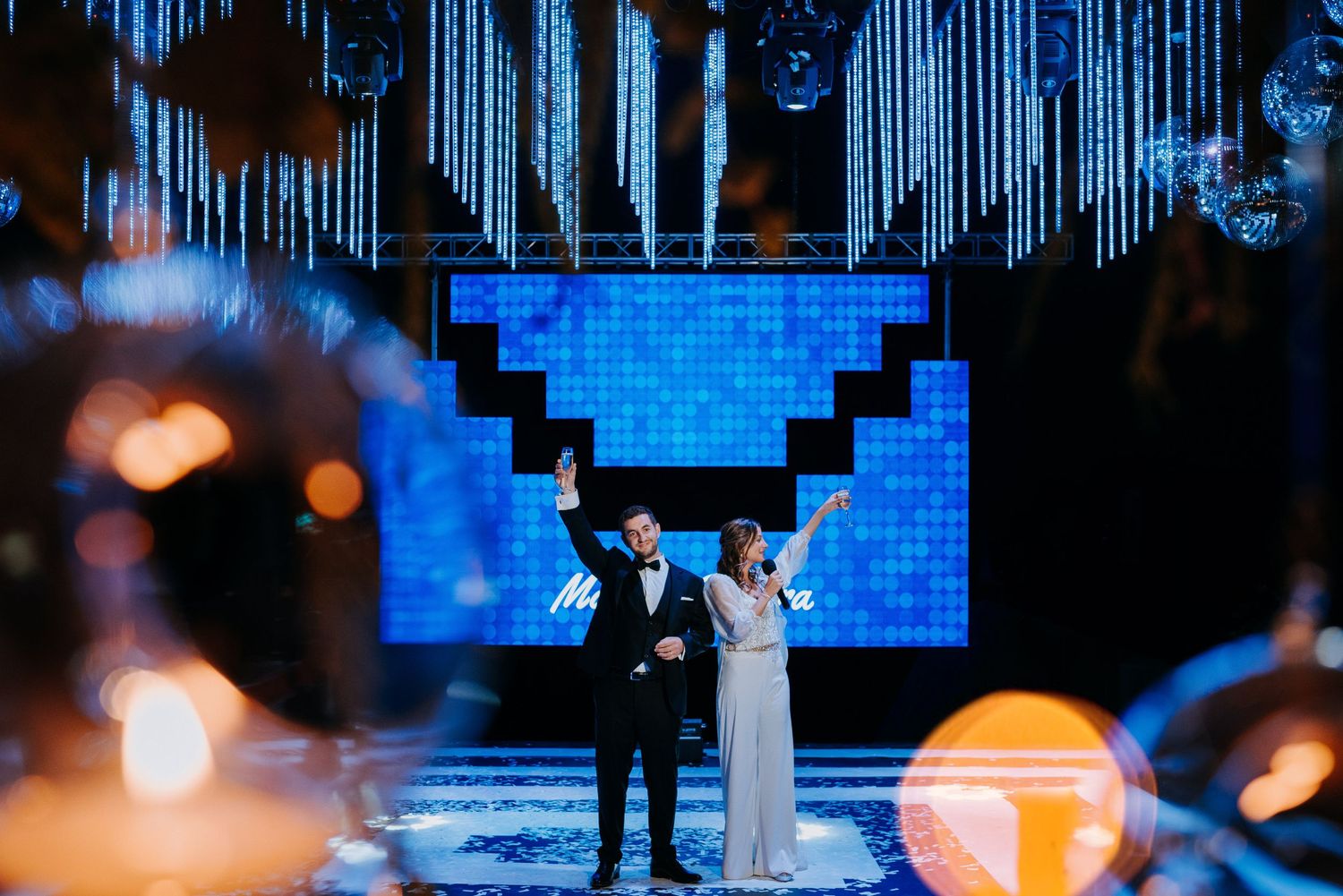 Couple celebrating at wedding reception on a stage with blue pixel art backdrop and hanging decor.