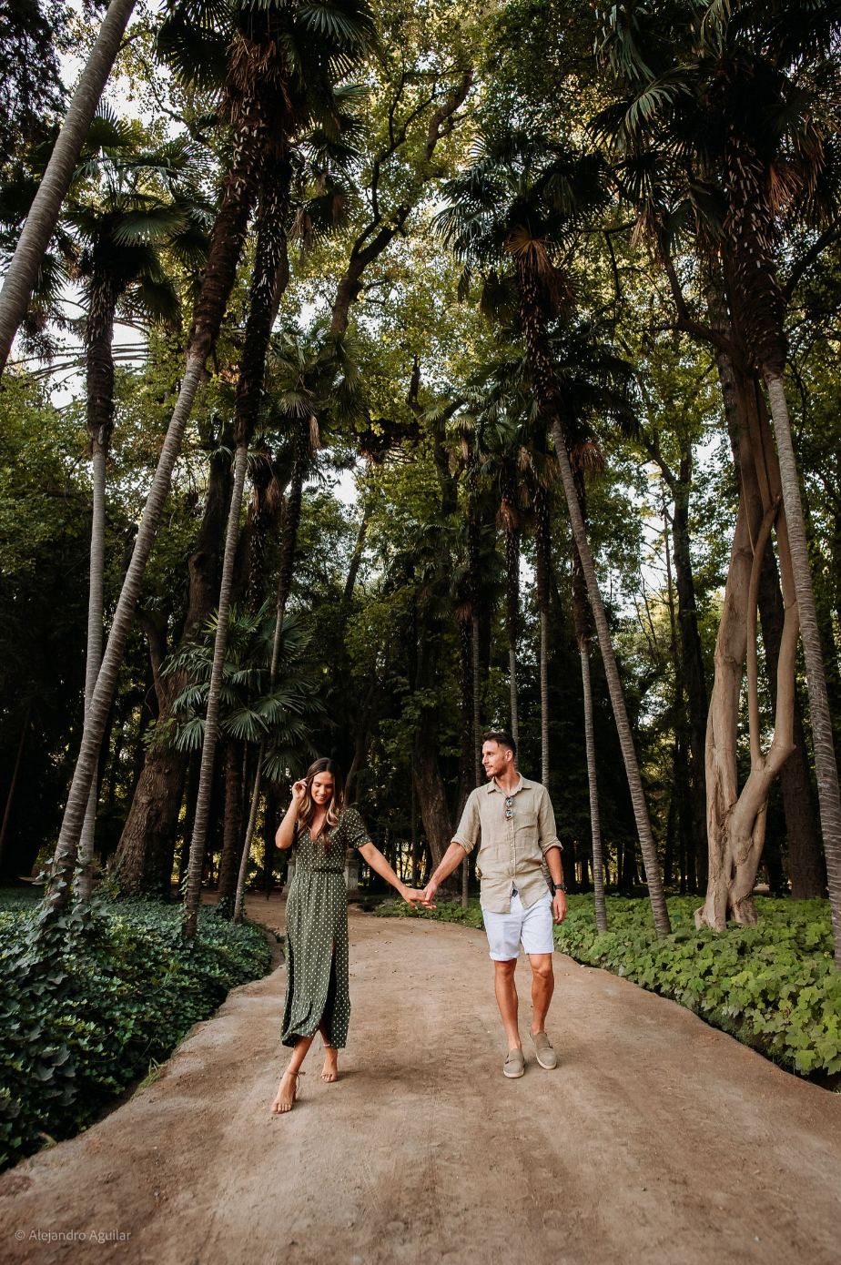 Couple holding hands while walking along a palm tree-lined path in a lush tropical garden.