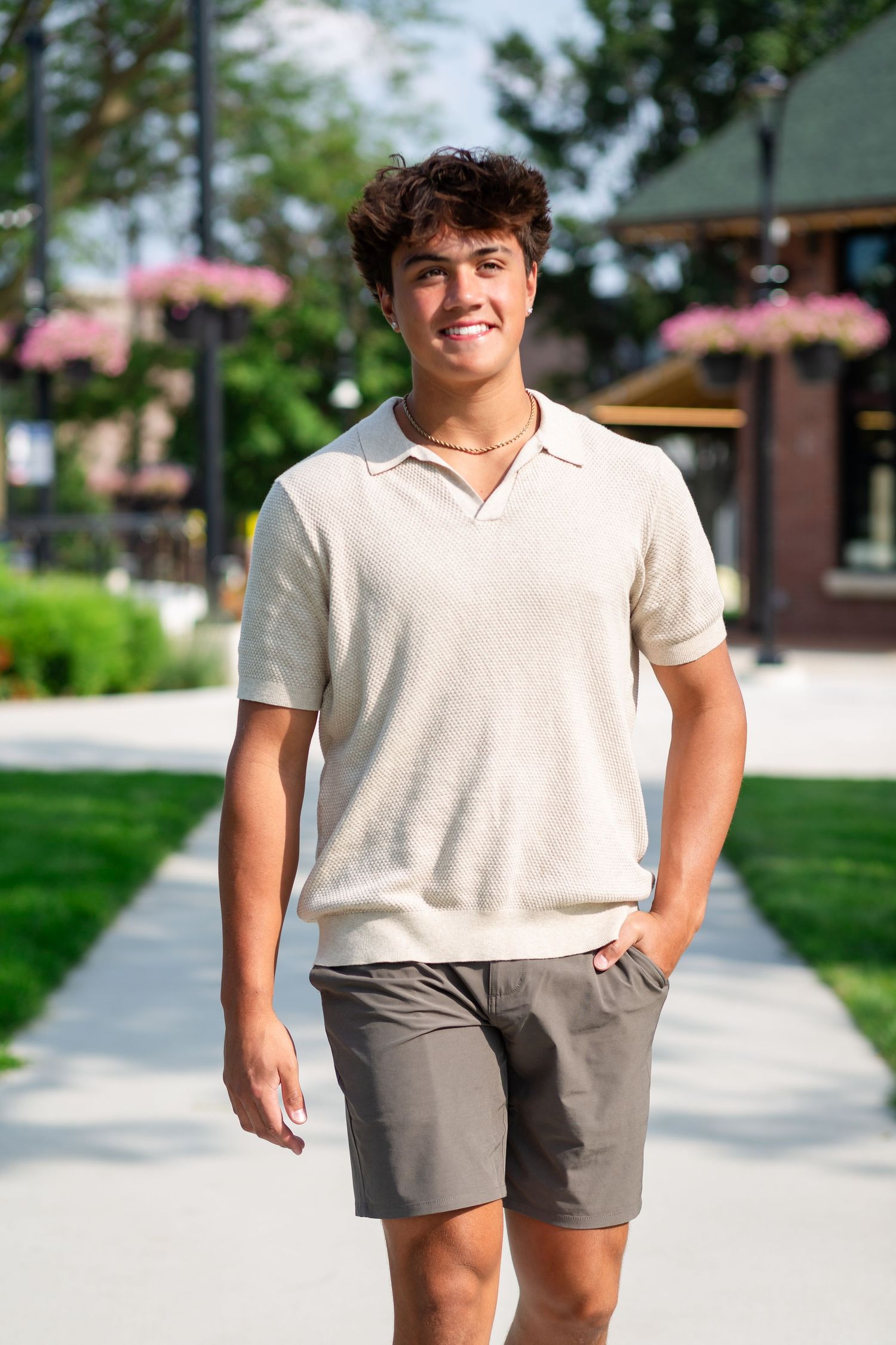 Young man in beige polo shirt and gray shorts smiling while walking on a sunny outdoor pathway.