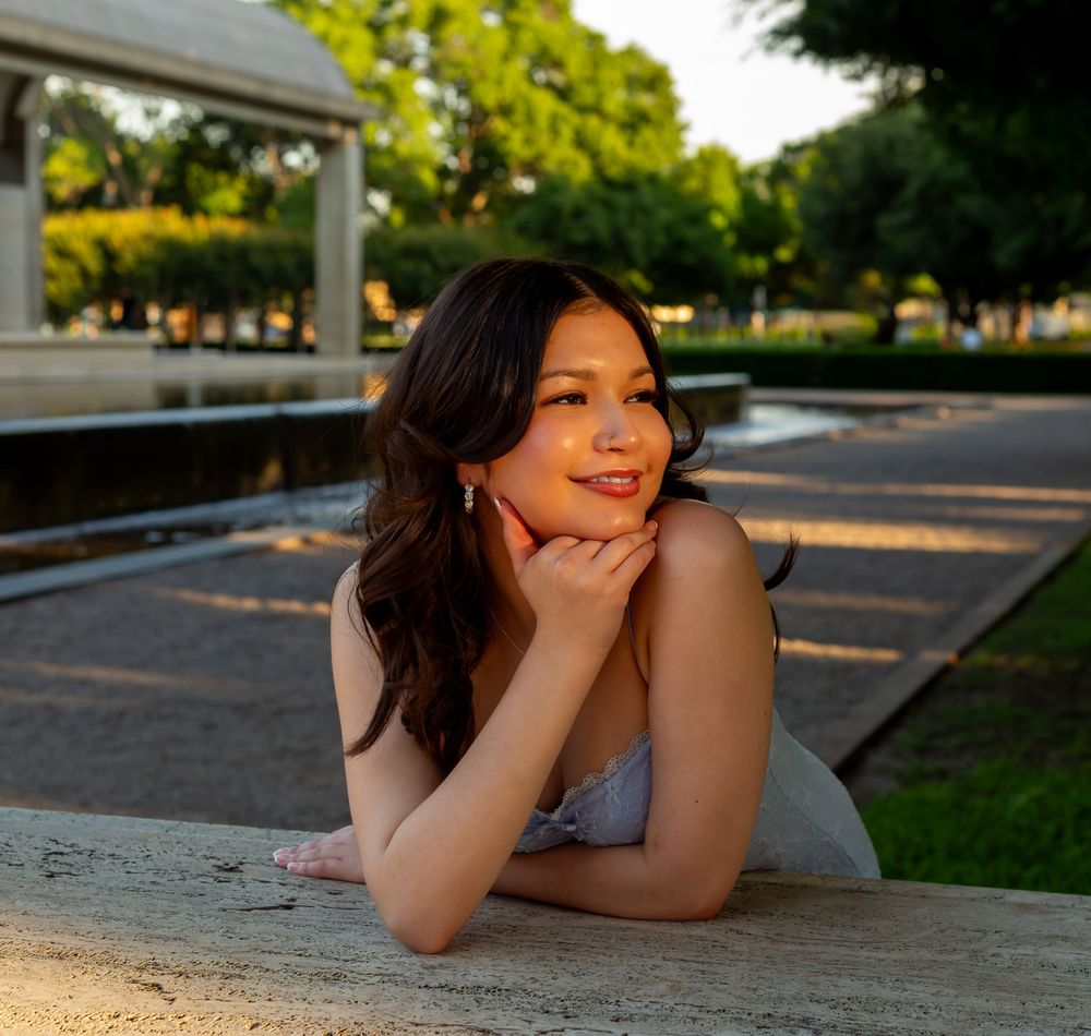 Young woman in white dress posing outdoors near a fountain at golden hour in a park.