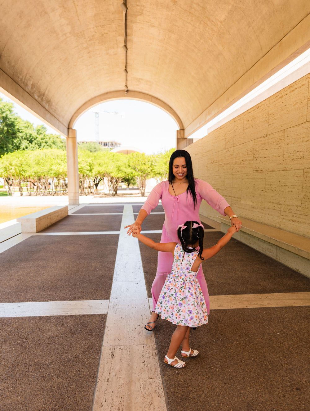 A mother in pink dances with her young daughter in a floral dress under an arched walkway outdoors.
