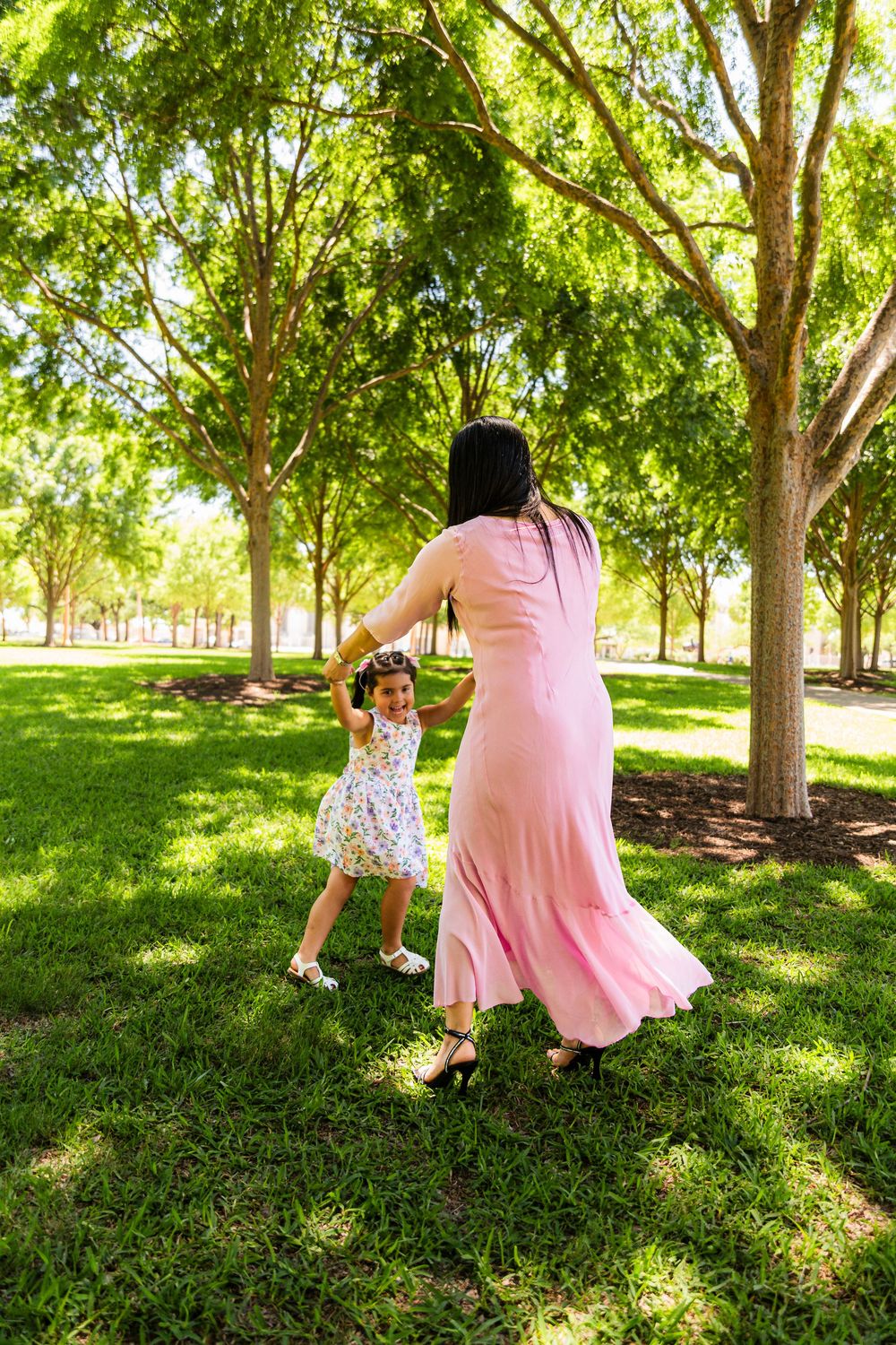 Mother and daughter dancing together in a sunny green park, wearing pink and floral dresses.