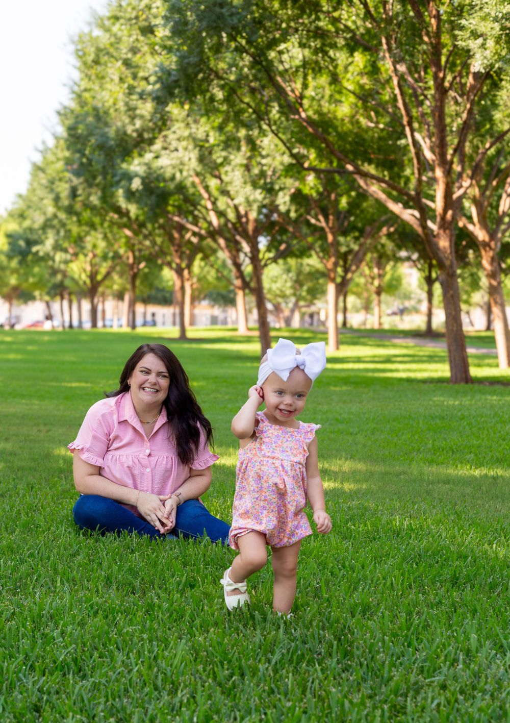 A mother in pink sits on green grass smiling as her toddler girl in a floral romper and bow walks nearby in a park.