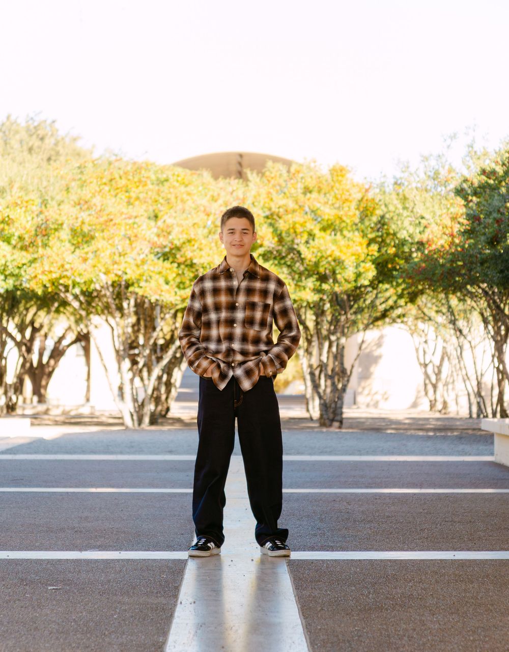 Young man in plaid shirt and black pants standing in parking lot with yellow-green trees in background.