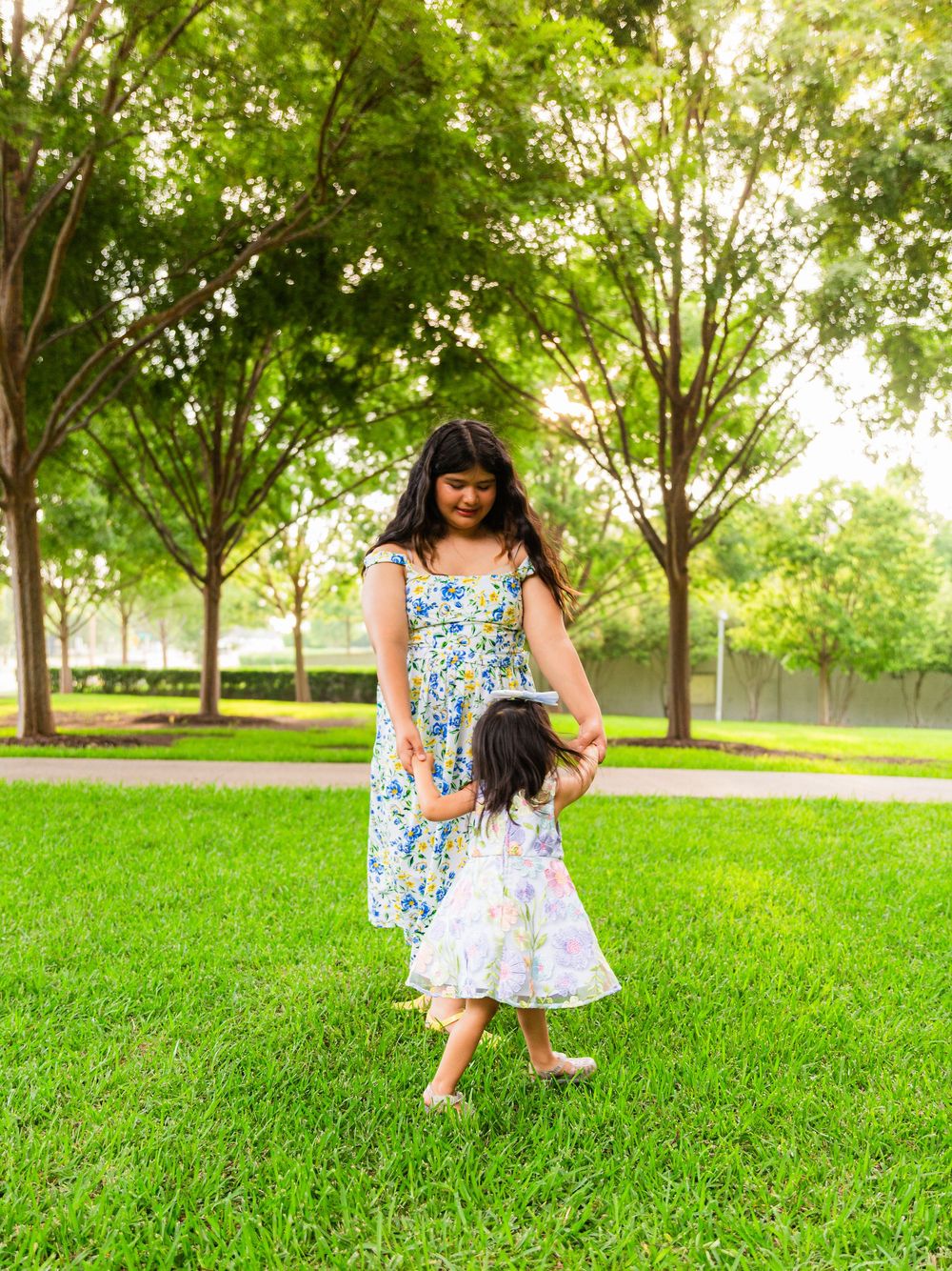 A mother and young daughter in floral dresses hold hands while walking on green grass in a sunny park.