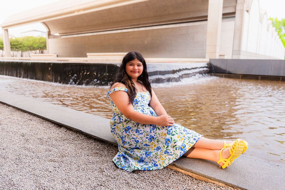 Pregnant woman in floral dress and yellow heels sitting by urban fountain under highway overpass.