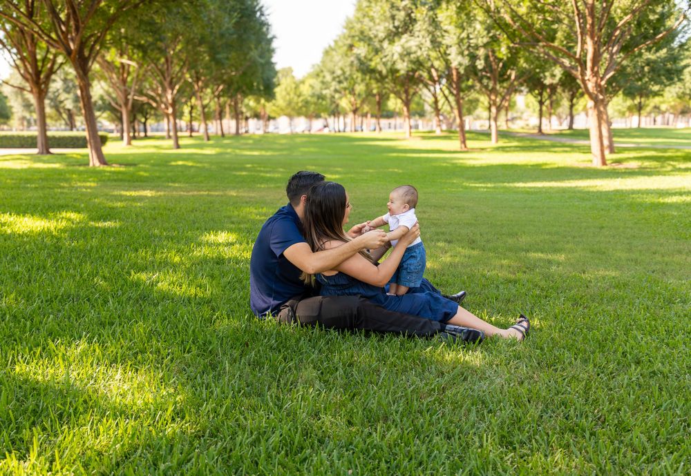 A mother sits on lush green grass in a sunny park, lovingly holding her baby up in the air.