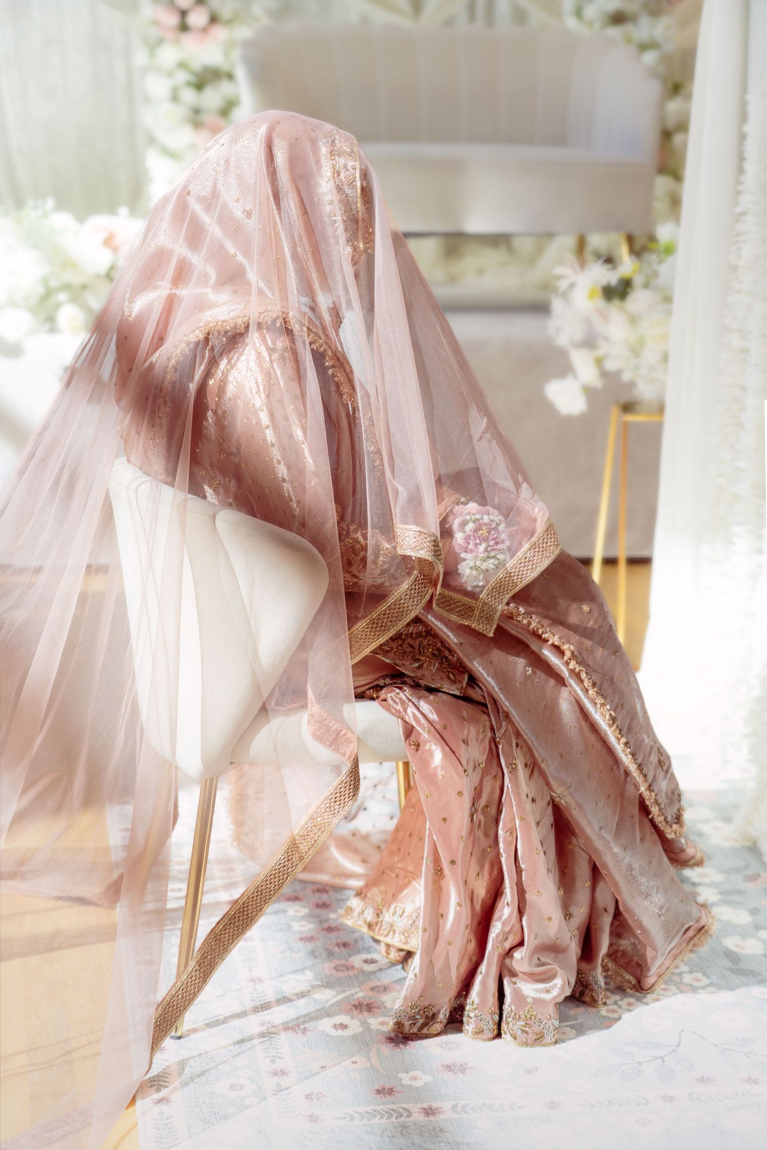 Bride in blush pink embroidered lehenga with sheer veil draped over head, seated at elegant floral wedding ceremony.