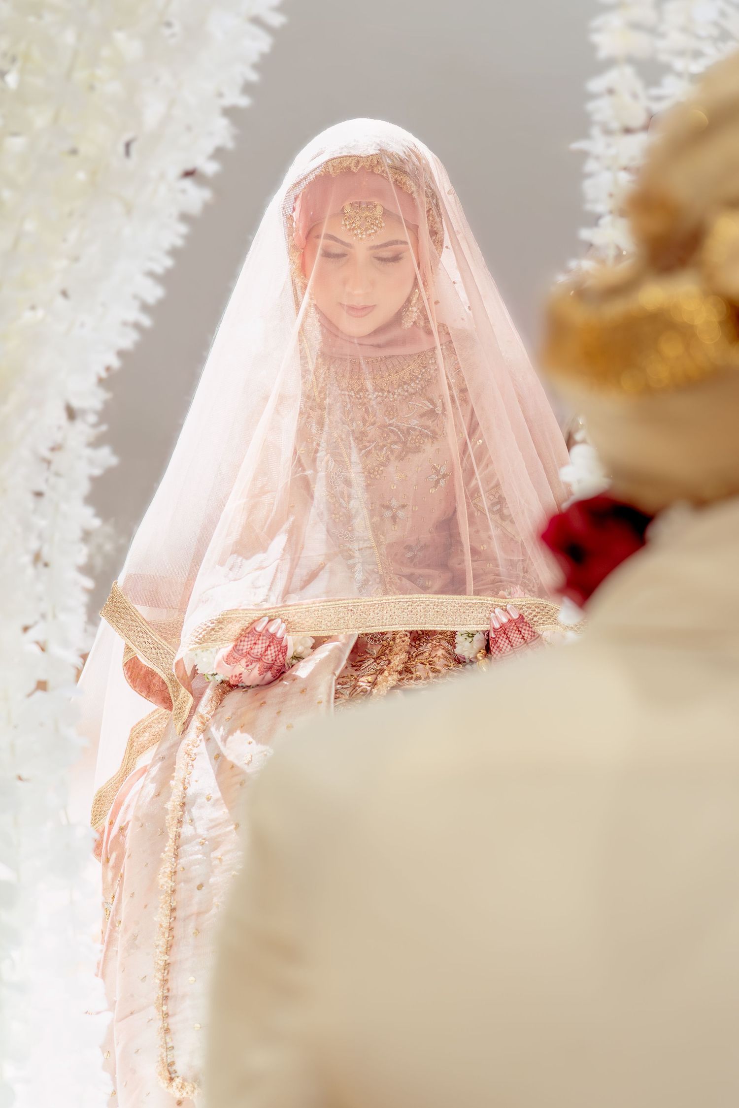 A bride in traditional South Asian wedding attire with gold jewelry and a delicate pink veil during a ceremony.
