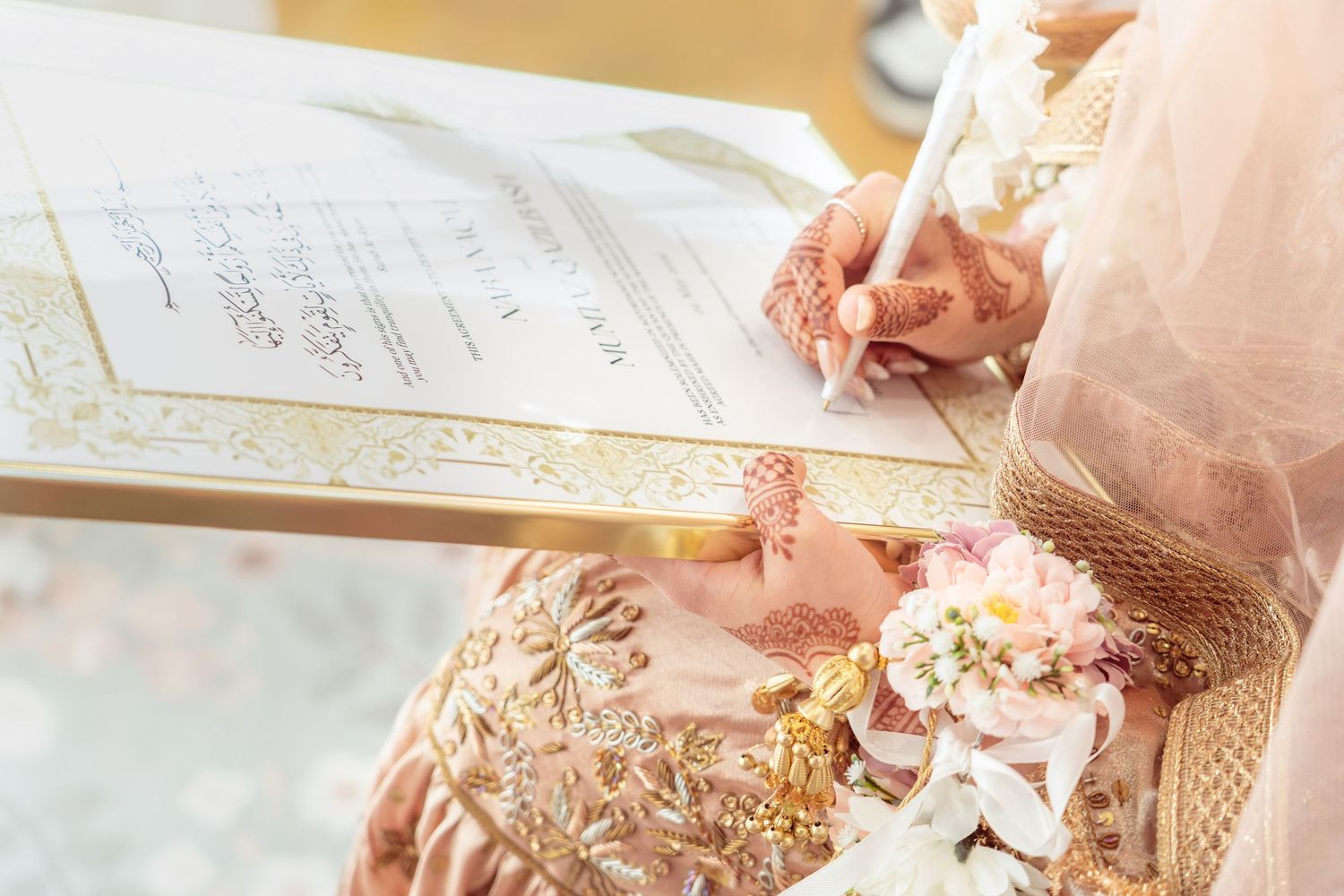 Bride in gold embroidered dress signing wedding nikah certificate with floral bouquet during Islamic marriage ceremony.