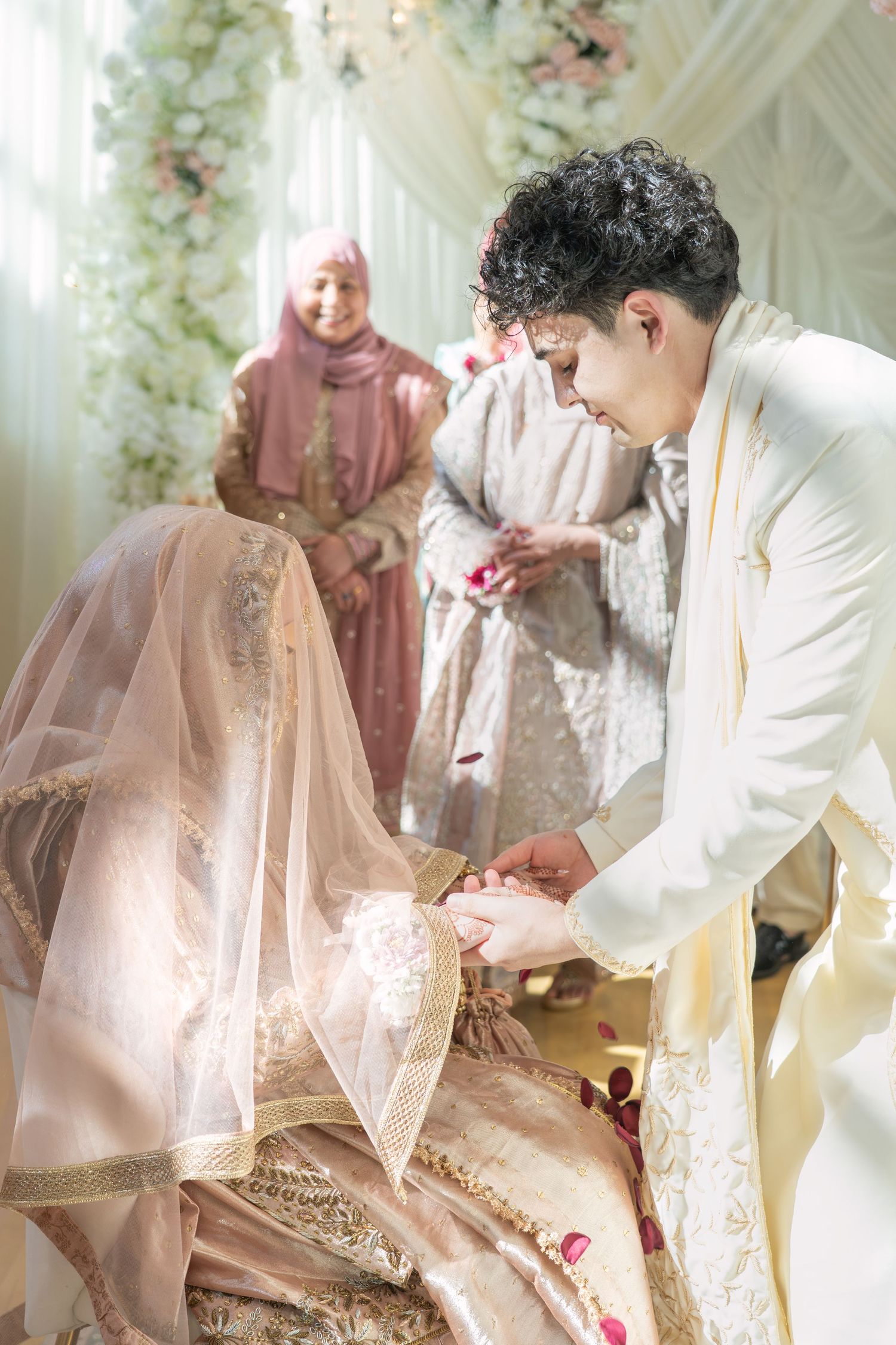 Groom in white suit places ring on bride's hand during Islamic wedding ceremony with guests in background.
