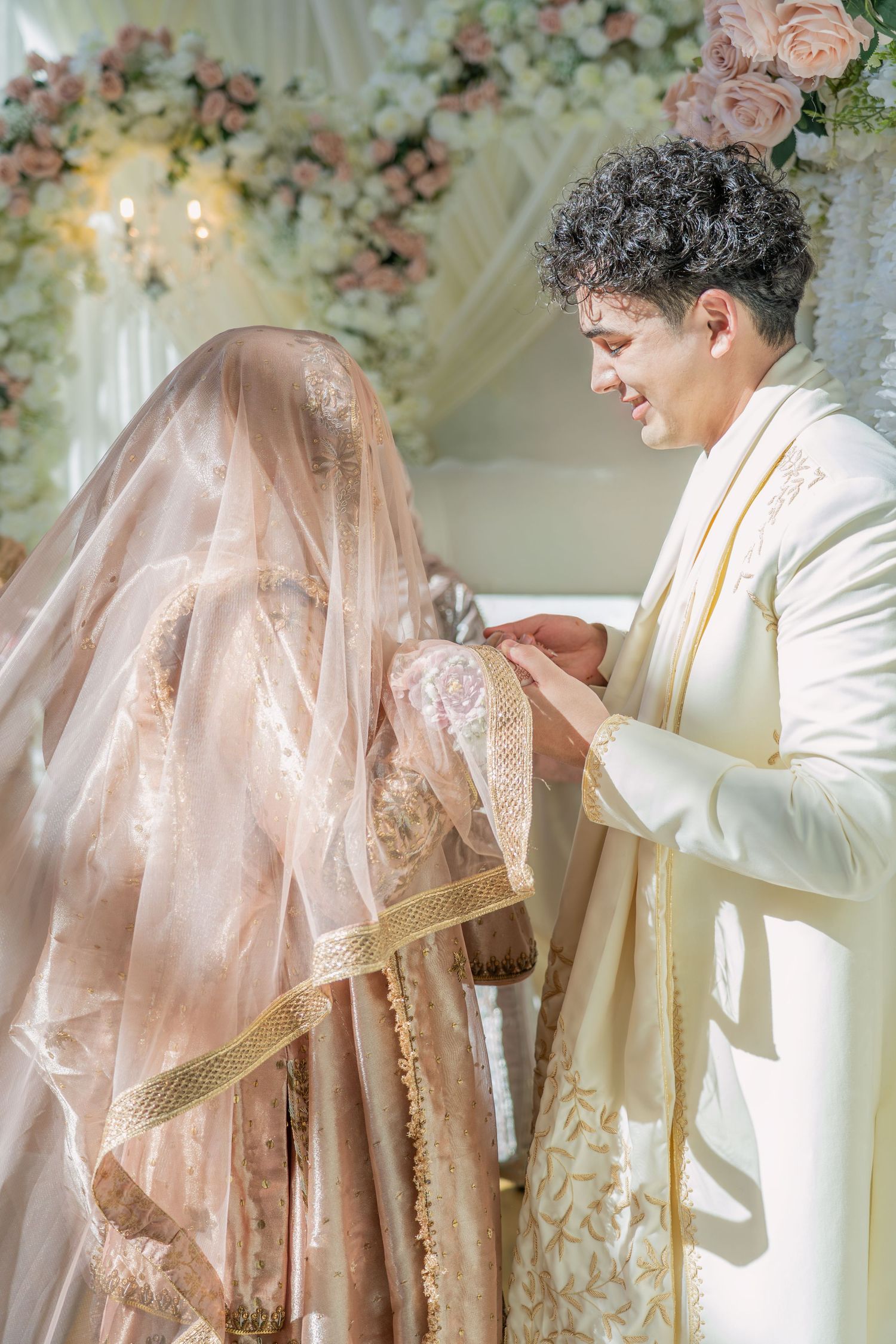 A groom in white sherwani holds hands with his veiled bride during a traditional South Asian wedding ceremony.