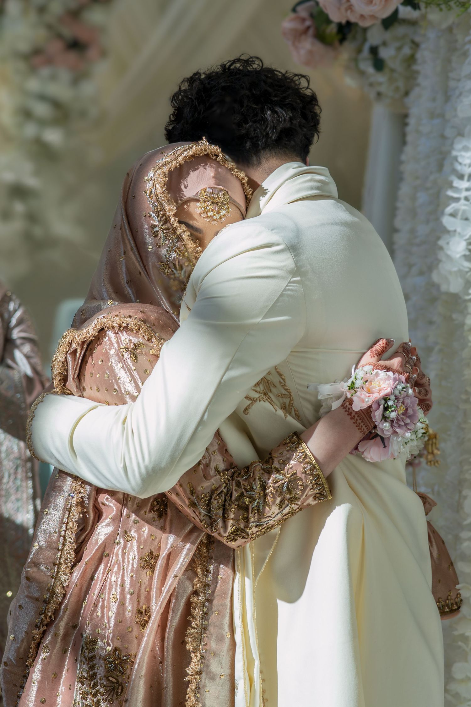 A bride and groom in traditional South Asian wedding attire share a warm embrace, with floral decorations in the background.